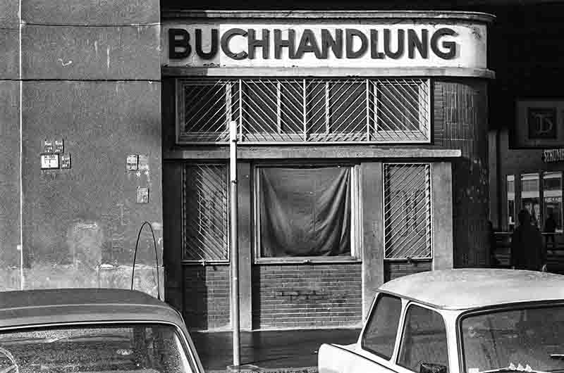 A black and white photograph of a vintage German bookshop named 'BUCHHANDLUNG' with heavy metal security bars over the windows. A fabric curtain covers the main display window. In the foreground, the roofs and windows of two vintage European cars, including a Trabant, are visible parked on the street.
