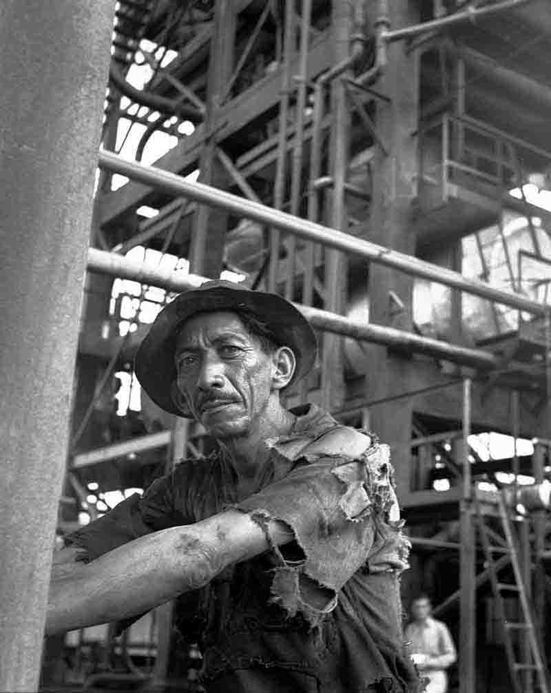 Vintage print of a man with a haggard yet proud expression standing in a torn shirt on an oil platform