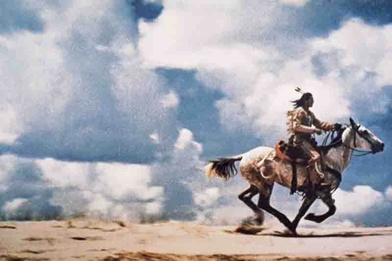 Conceptual Photography A full-shot image shows a person with long, dark hair wearing a headdress with a single feather, riding a white horse at a gallop across a sandy, barren landscape. The person is in profile, facing toward the right side of the image. The horse is kicking up a cloud of dust, and the sky above is filled with dramatic white and gray clouds.