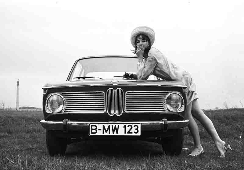 Monochrome photograph of a woman leaning on the hood of a vintage BMW car, embodying a sense of ease and style in a calm and inviting atmosphere.