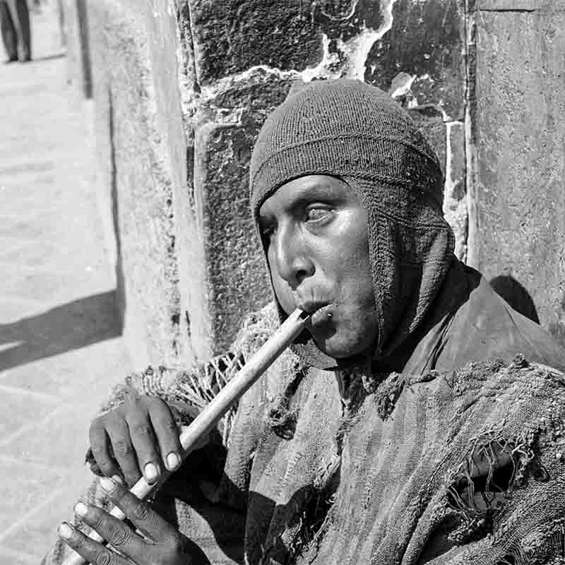 A close-up, black and white limited edition photo shows a man sitting and playing a long, slender flute. He is wearing a knitted cap pulled low over his forehead and a rough, textured garment.