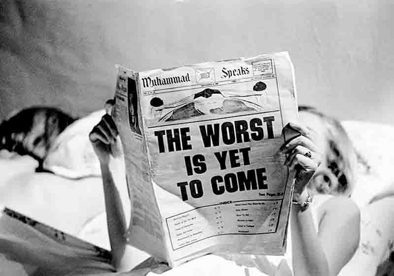 A black and white photograph by Steve Schapiro titled 'Reflection of Zeitgeist.'' The image shows a woman lying in bed, her face mostly obscured by a newspaper she is holding up in front of her. Only her eyes, hands, and blonde hair are partially visible.