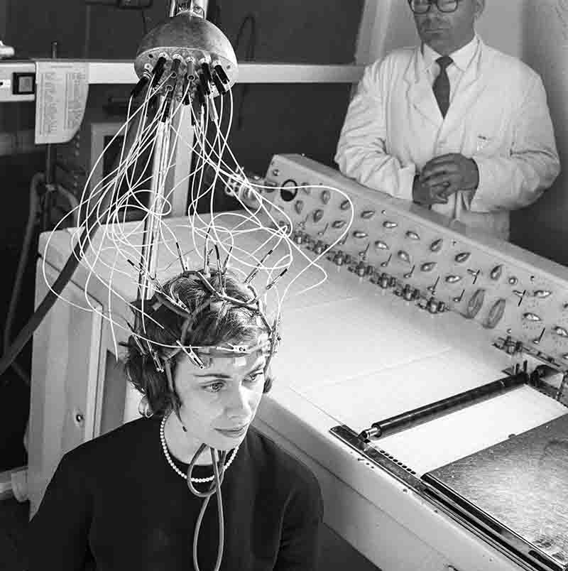 A woman is positioned in front of a machine, wires attached to her head, illustrating a scenario involving advanced technology.