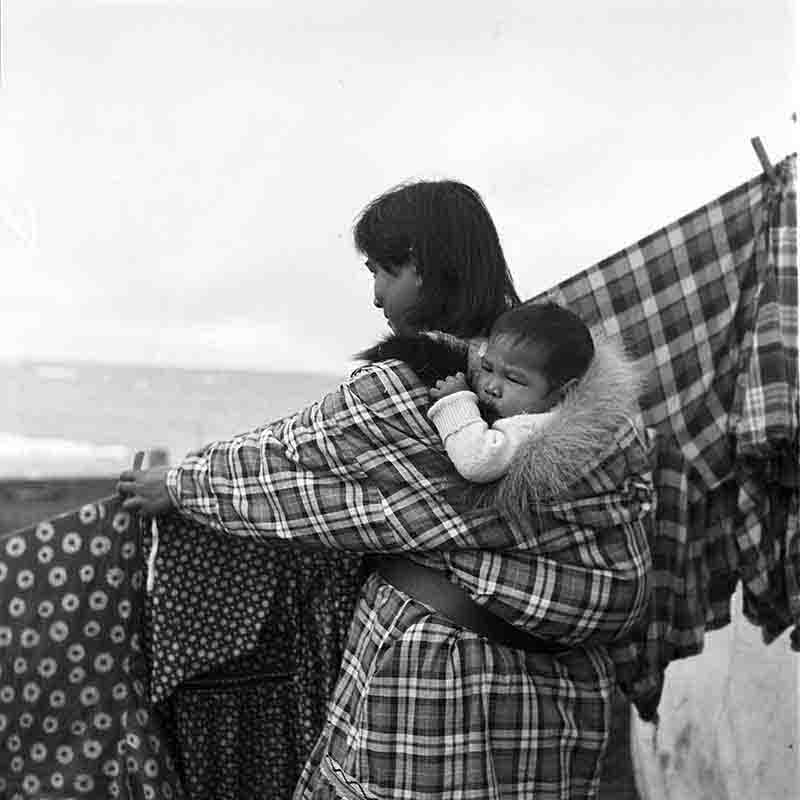 An Inuid woman hangs up laundry while carrying her baby on the traditional child carrier bag.