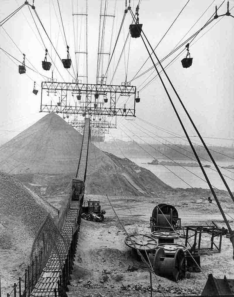 A monochrome vintage photograph depicting a cable car ascending a mountain, highlighting the contrast between the structure and the terrain.