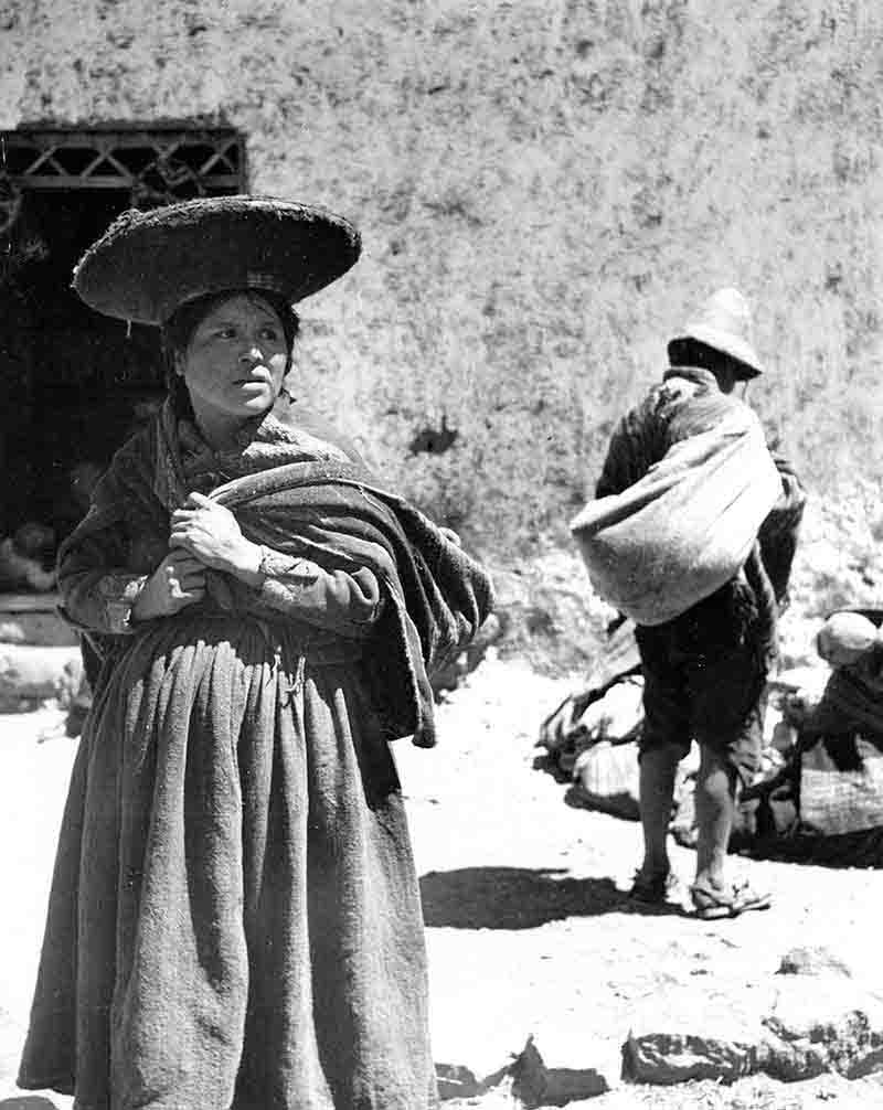 Vintage photograph of an indigenous woman from Peru carries a wicker basket atop her head, wih man in the background.